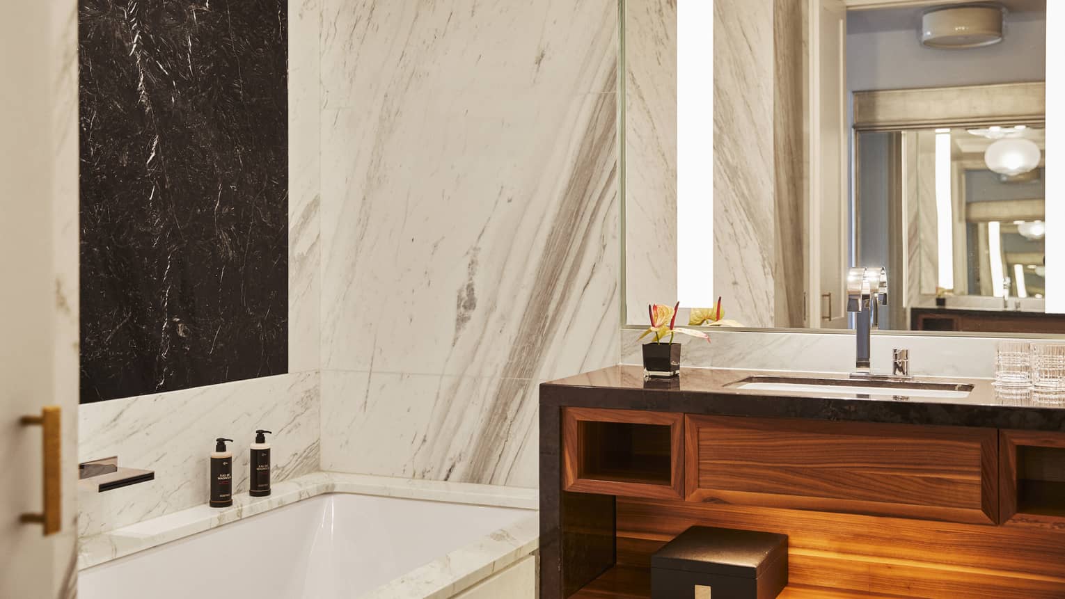 Luxury hotel bathroom with marble walls, black accent panel, built-in bathtub, wooden vanity and large mirror with lighting