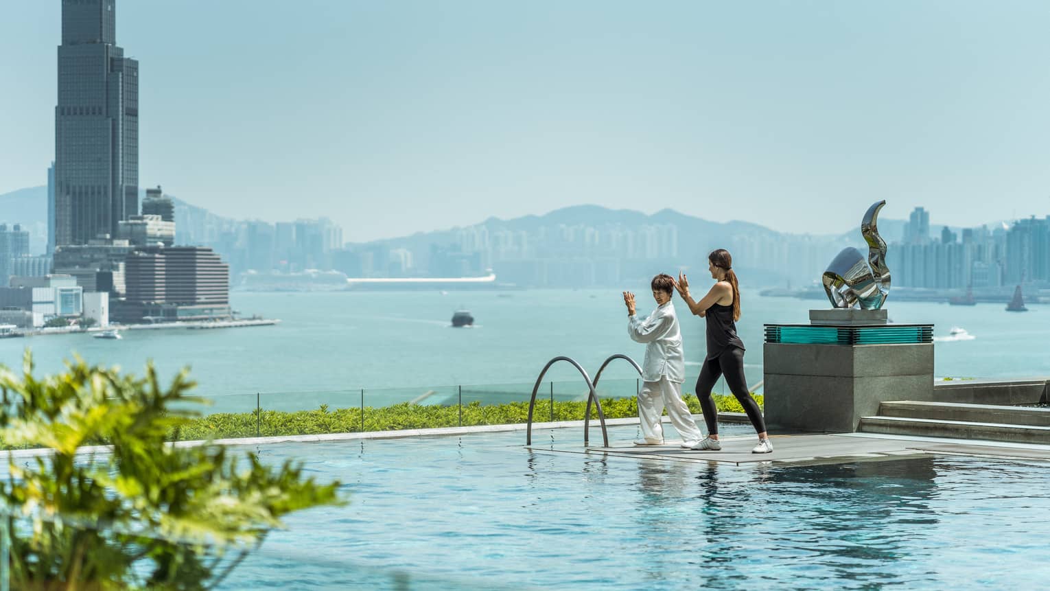 Two women in tai chi pose on platform on outdoor swimming pool overlooking Hong Kong waterfront