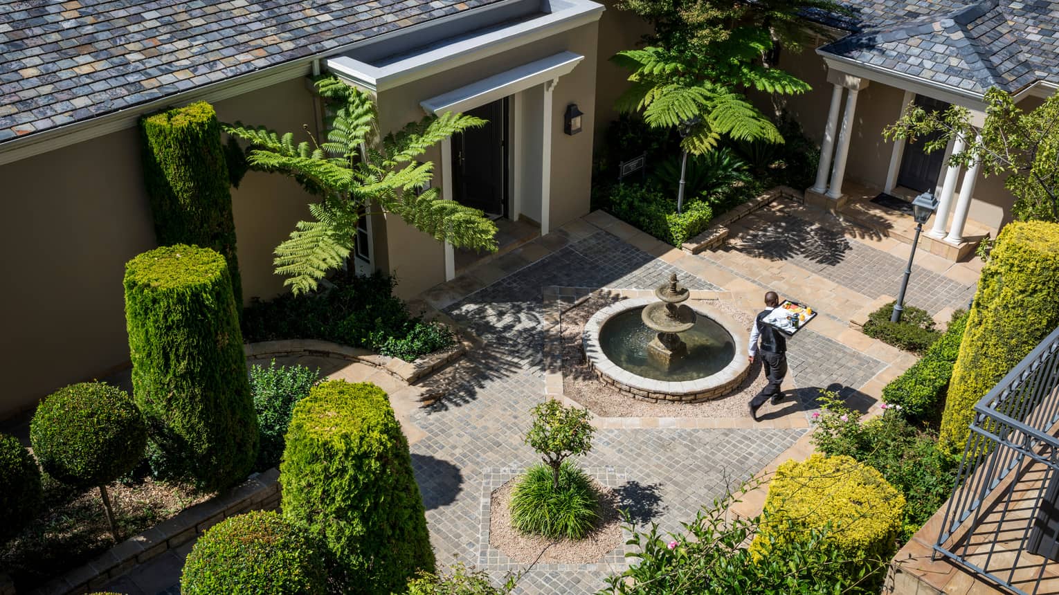Aerial view of waiter carrying tray through brick courtyard with manicured green shrubs