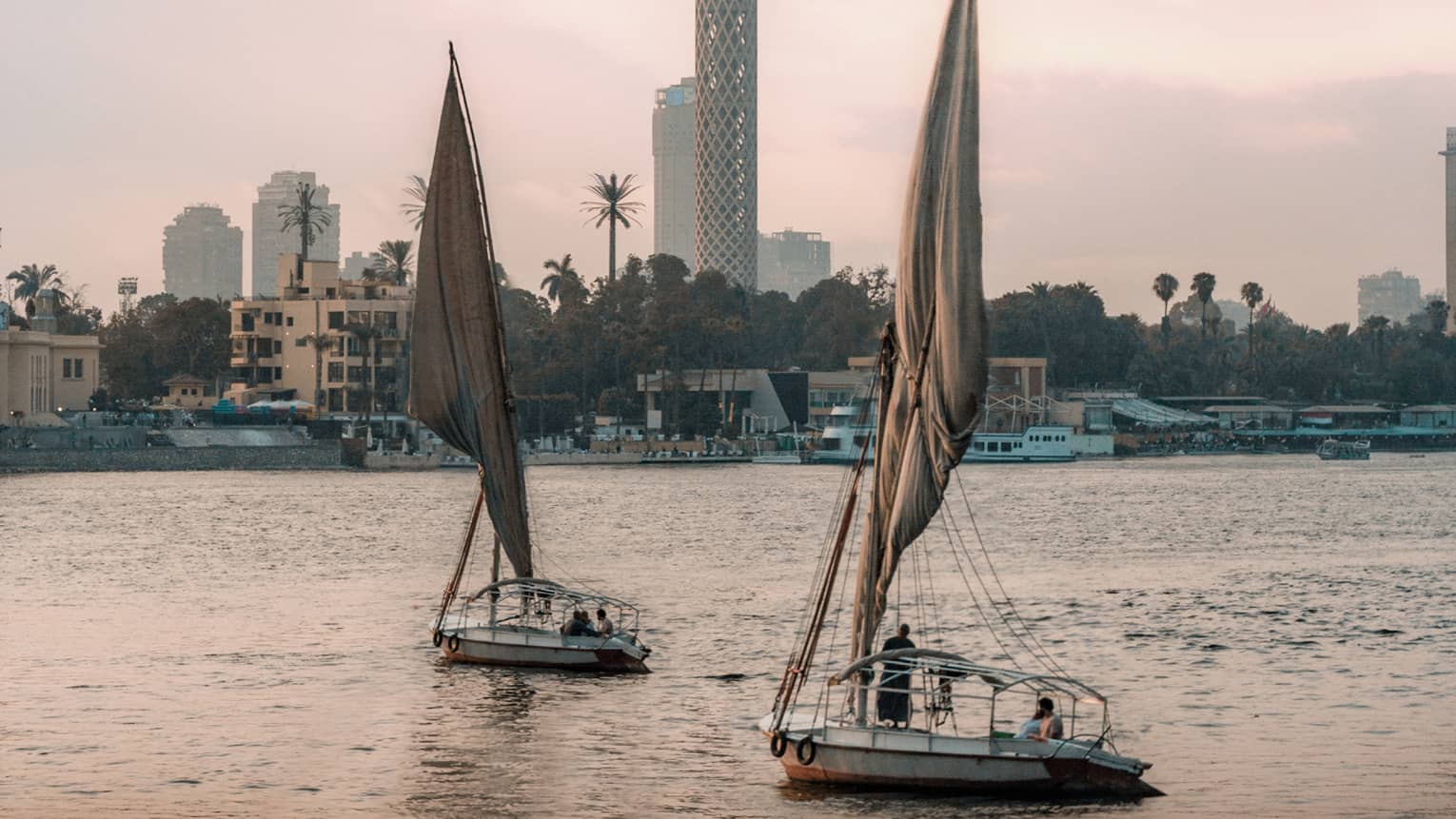Two felucca boats drift on the water in the evening light, a busy shoreline and Cairo Tower in the distance. 