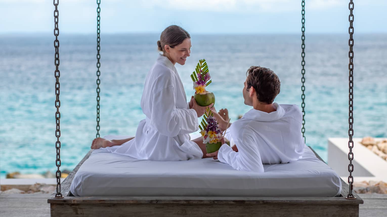 couple enjoying a sea view and a cocktail on a hanging beach bed whilst wearing white robes