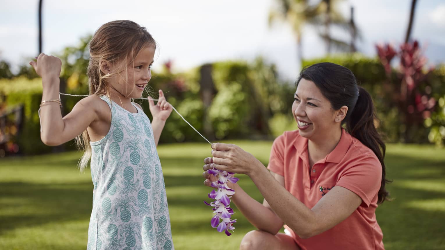 Girl in blue patterned dress smiling with staff member in orange polo, making lei