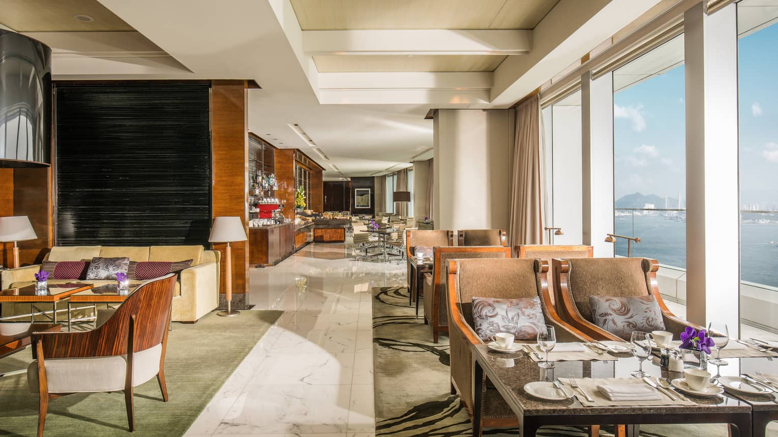Hotel lounge with modern wooden chairs and set dining tables lined up along tall windows with water view