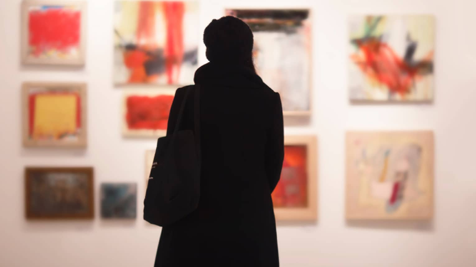 Silhouette of back of woman with hat bag looking at gallery wall with colourful art