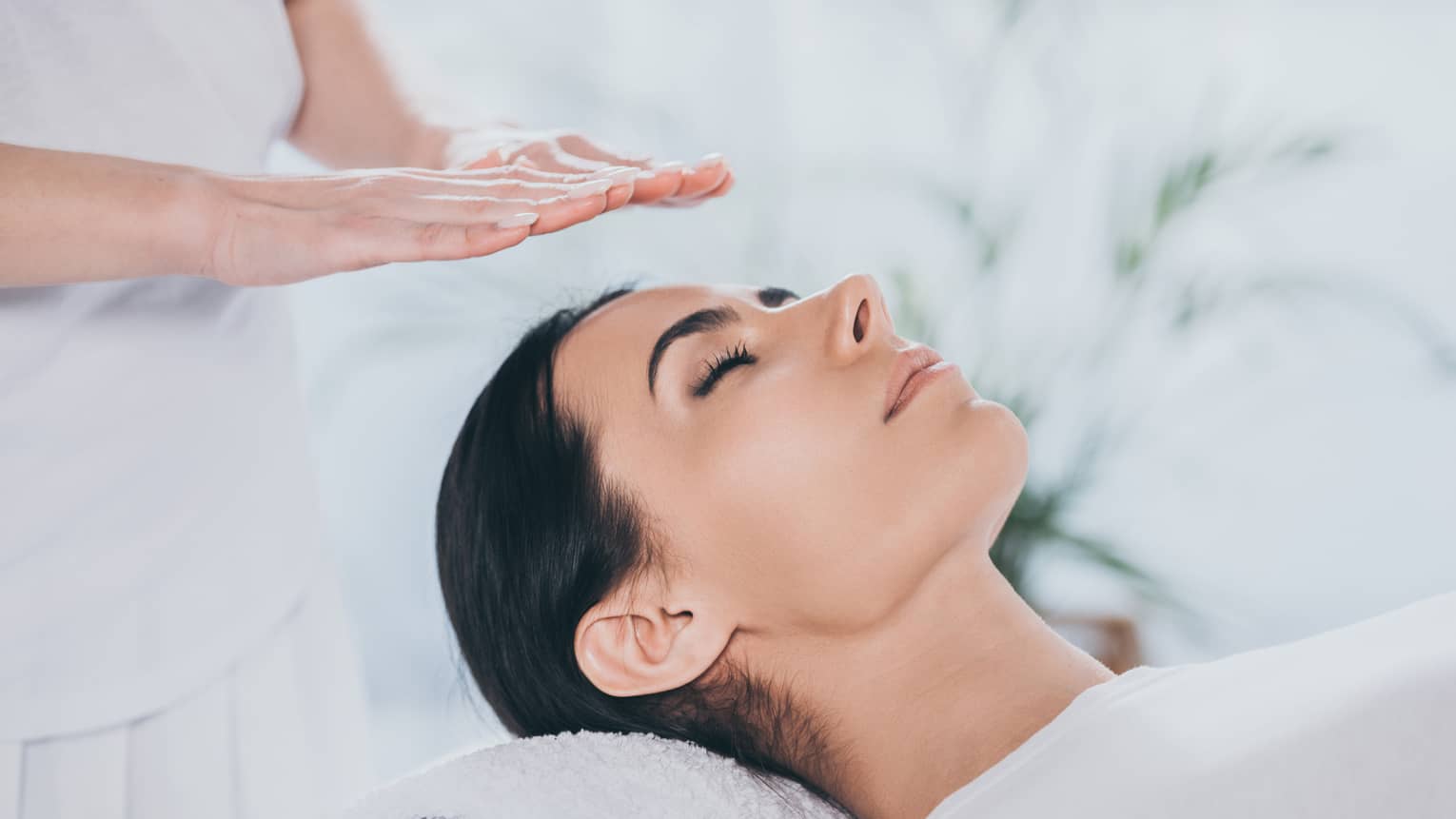 Woman with eyes closed lays on rolled up towel on spa table under therapists' hovering hands