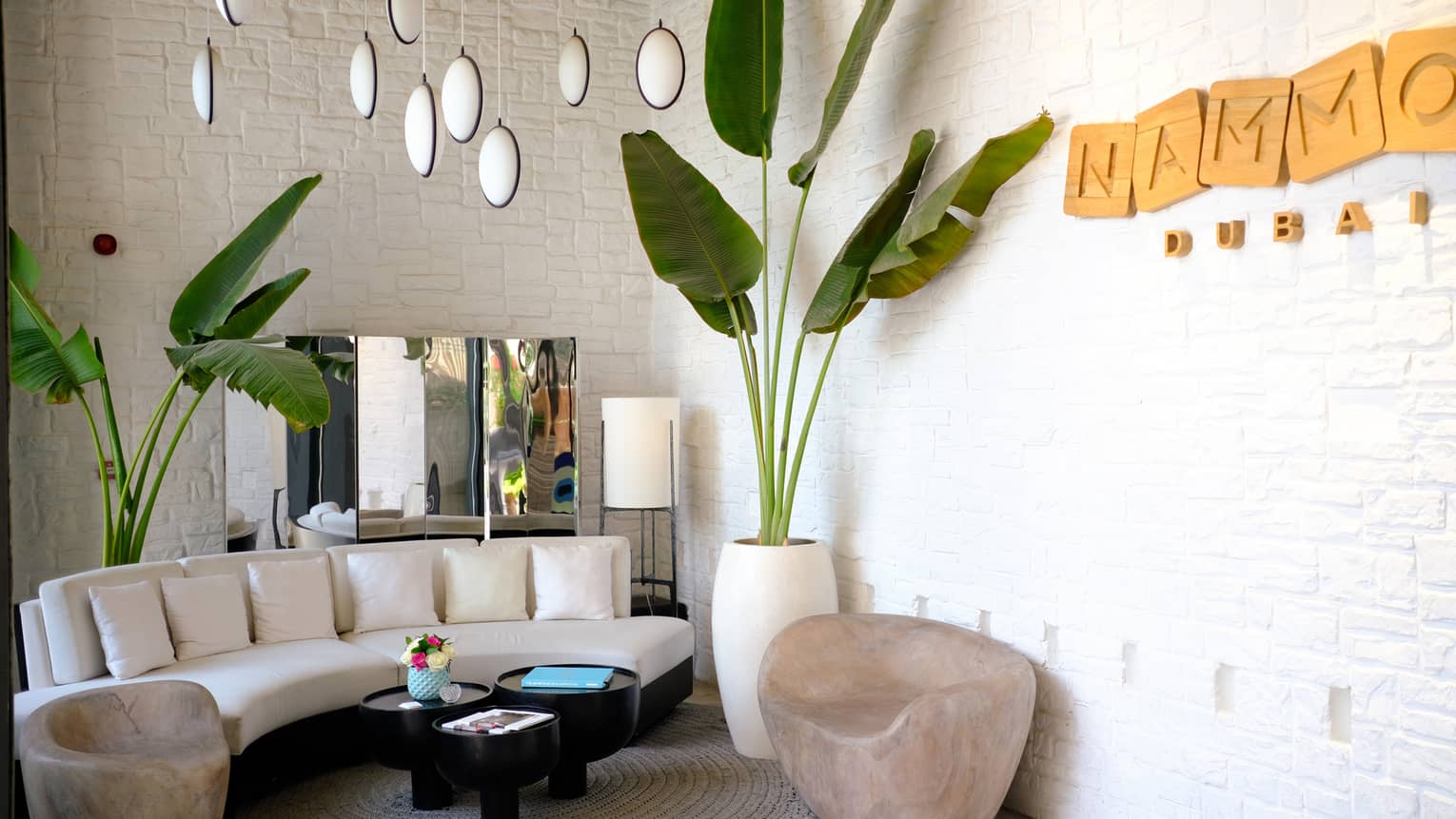 Indoor sitting area with curved white sofa, large palm plant, NAMMOS Dubai sign