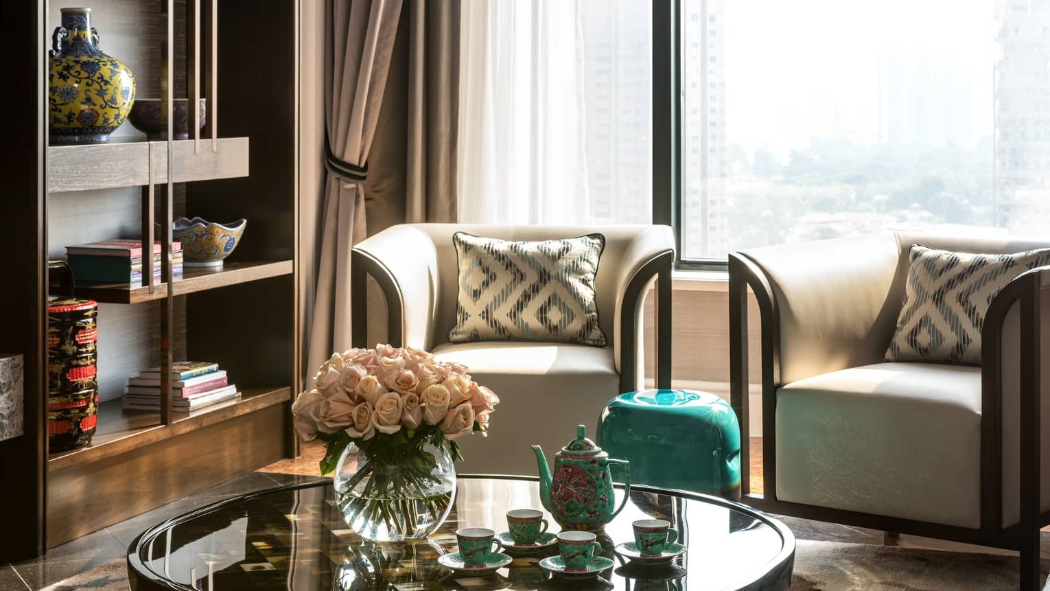 A cosy seating area with two armchairs, round coffee table holding a vase of pink roses and tea set, with a bookshelf displaying decorative vases and books nearby.