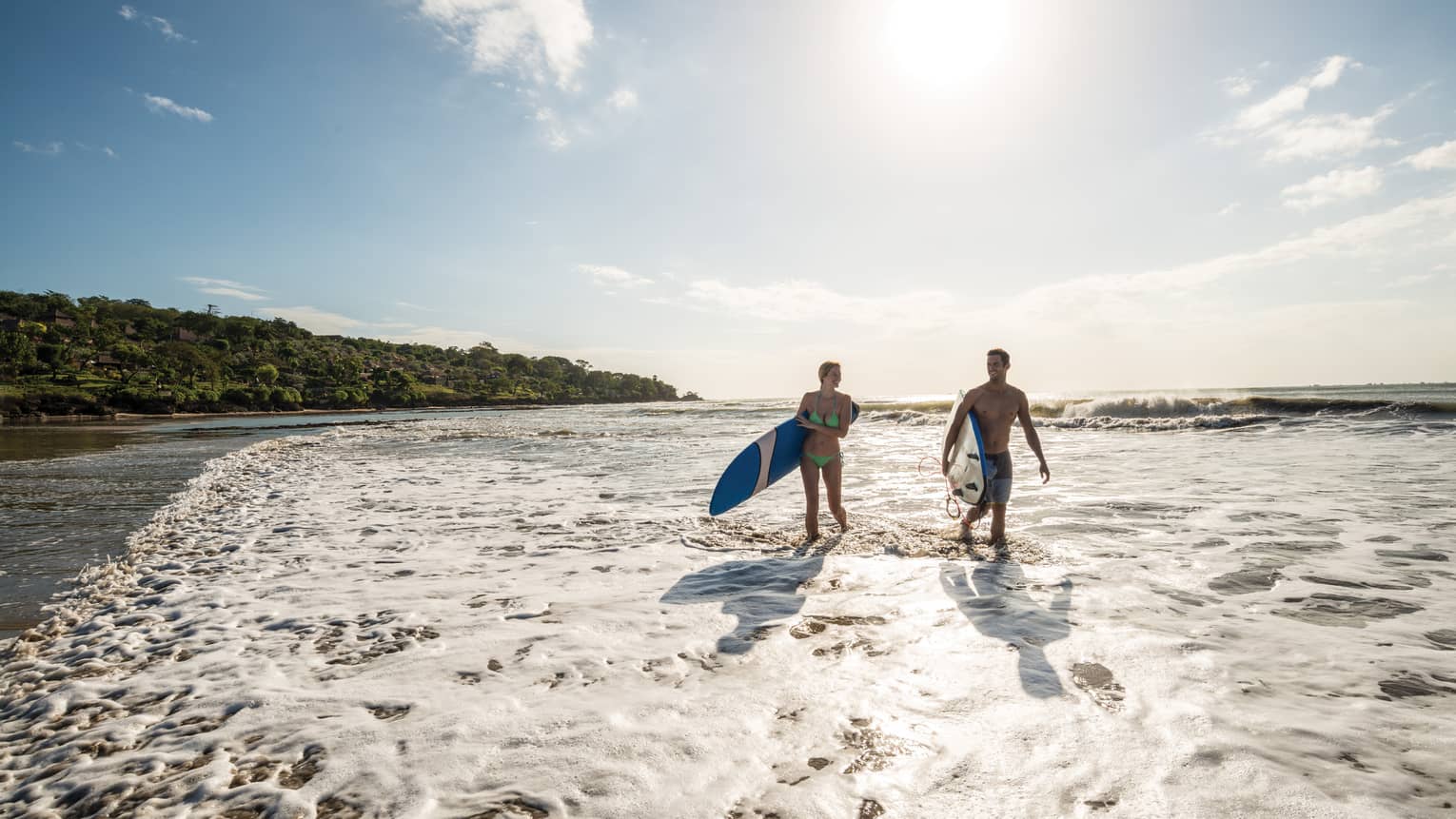 Silhouettes of man and woman in swimsuits, carrying surfboards up beach through tide at sunset