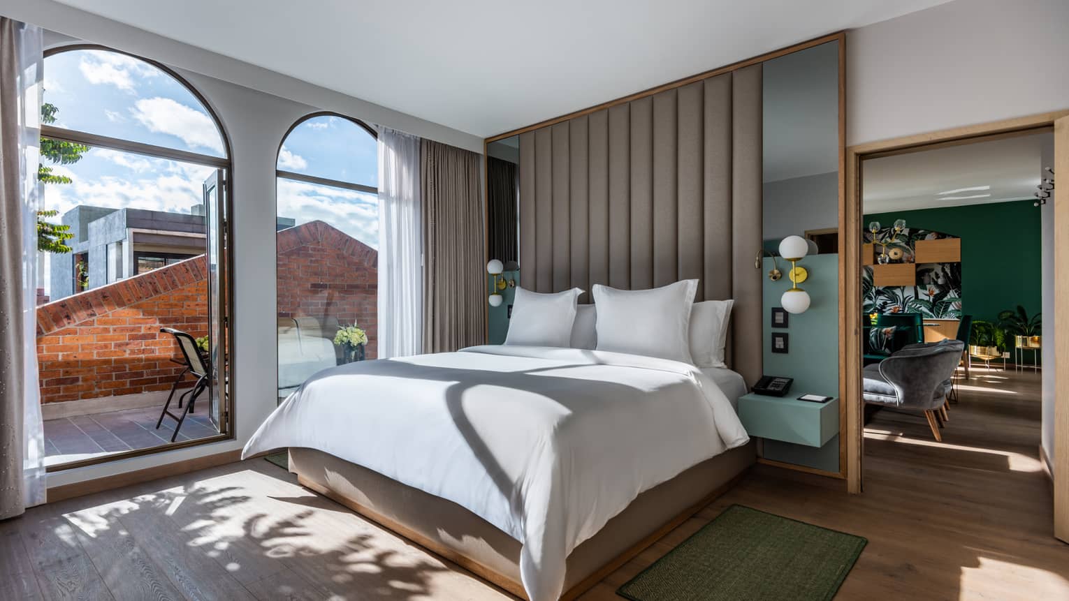 Guest room with bed and white linens, floor-to-ceiling windows overlooking the terrace, living/dining area just visible around the partition