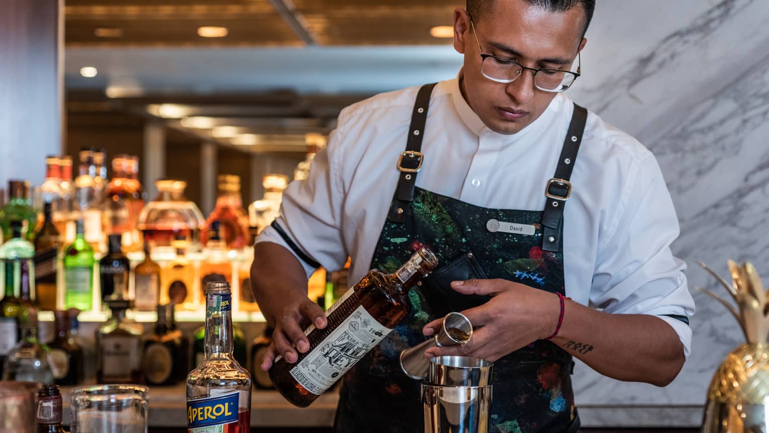 A bartender in a floral apron pours a brown liquor into a silver mixing glass.