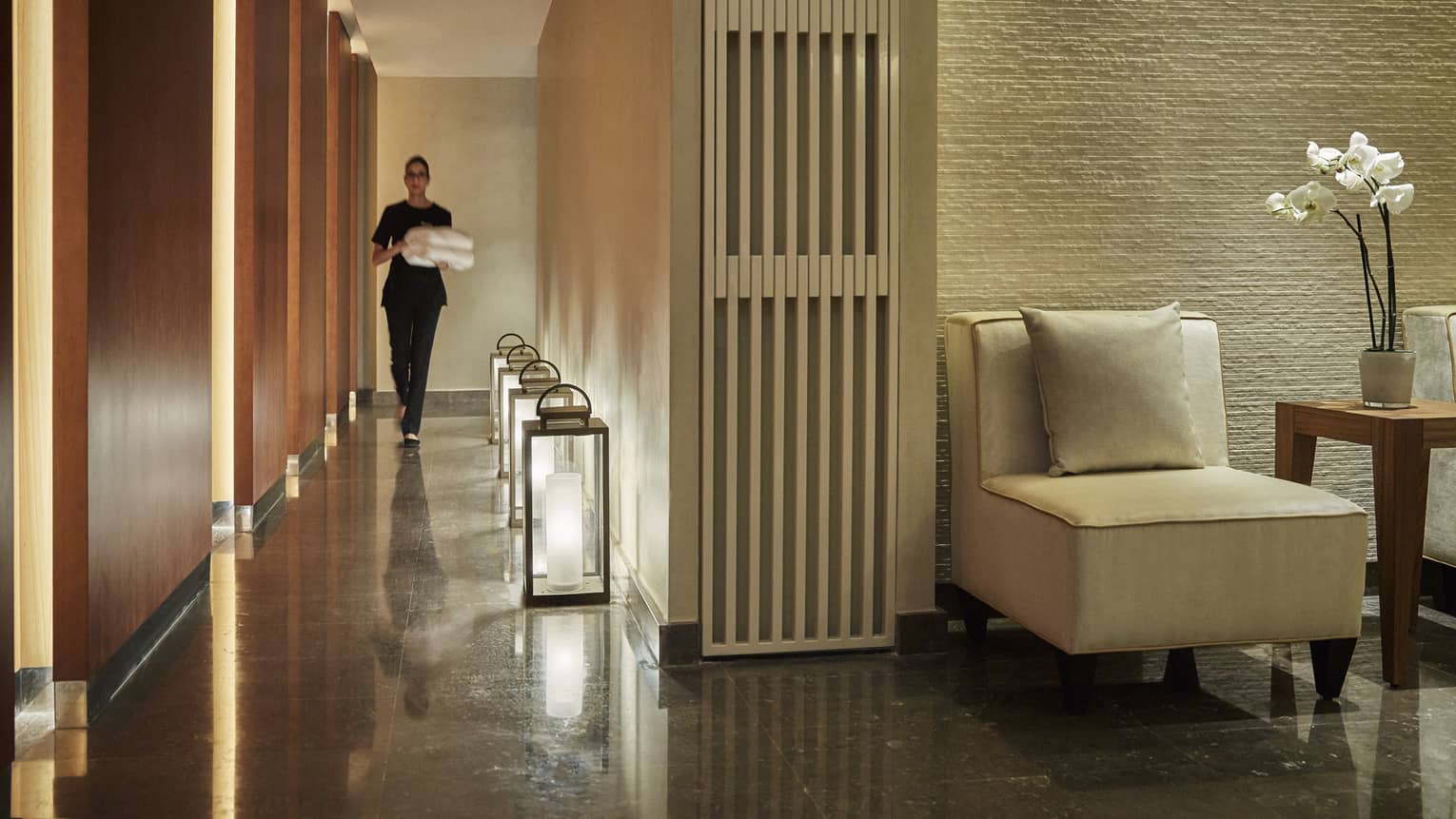 Woman carrying folded white towels walks down Le Spa hall past large lanterns along floor
