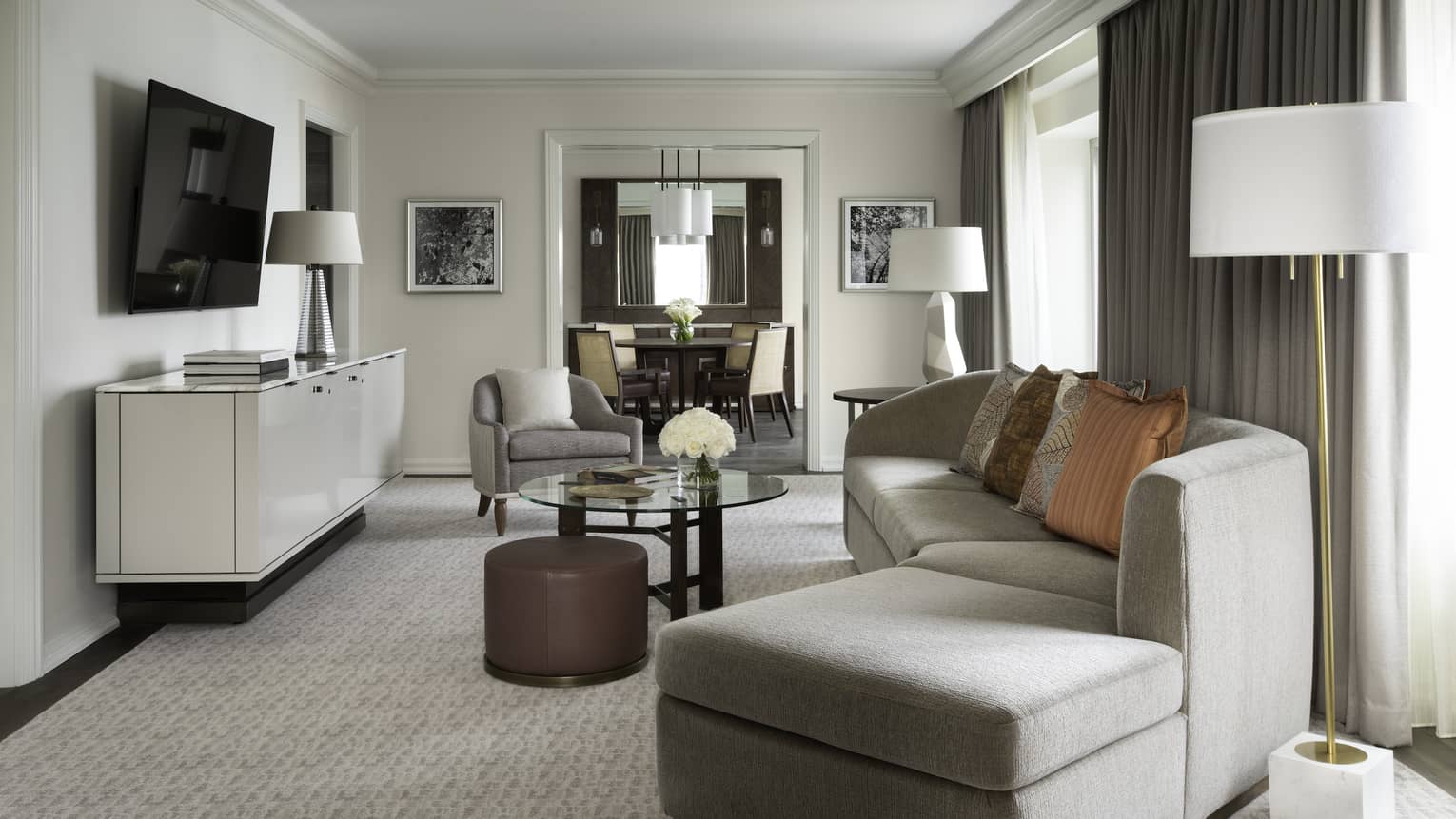 Living area with grey curved sofa, glass table, beige carpet, TV on wall, curtains