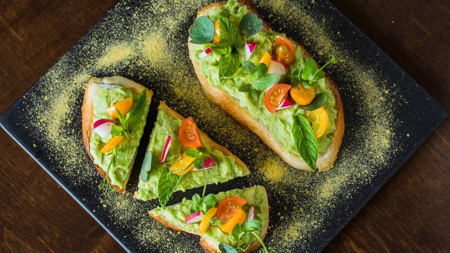 Toast topped with avocado, tomatoes and herbs, served on a black platter.