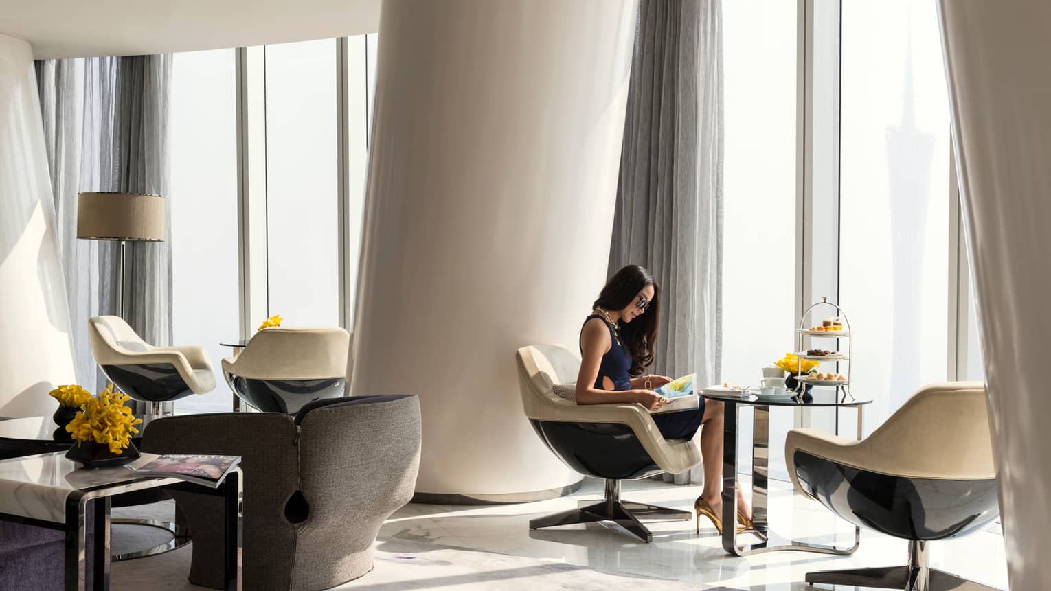 A guest sits in a neutral-toned room with a panoramic view and curved, modern design, a tiered tray of colourful fare nearby.
