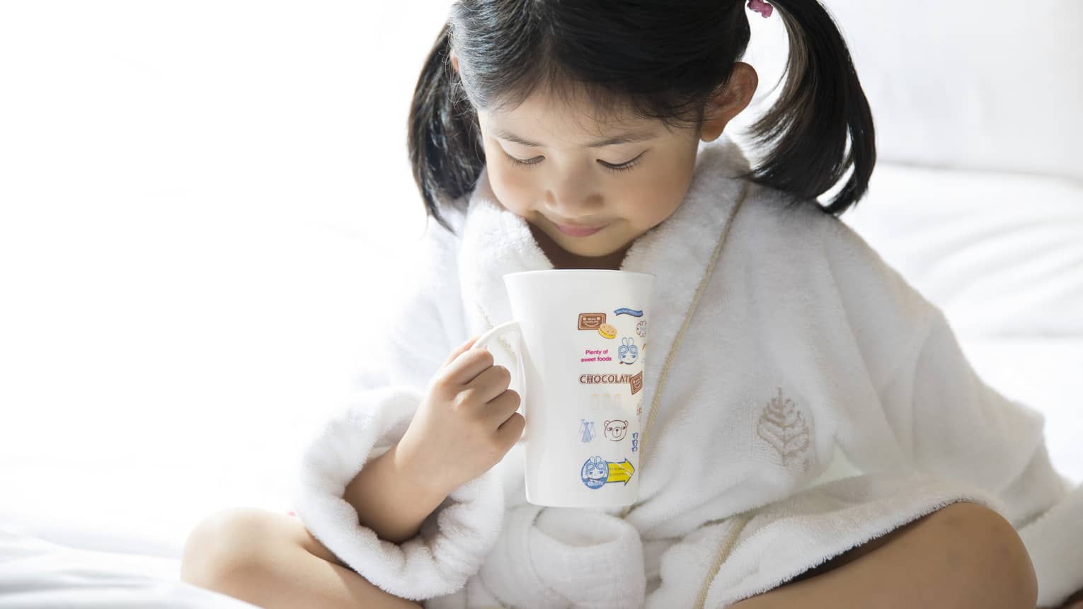 Young child in white bathrobe with hair in ponytails sits cross-legged on bed, holds mug of hot chocolate