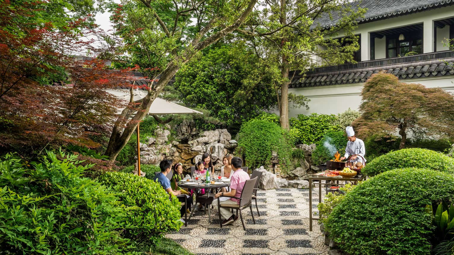 Family dines at round patio table by chef at barbecue in garden
