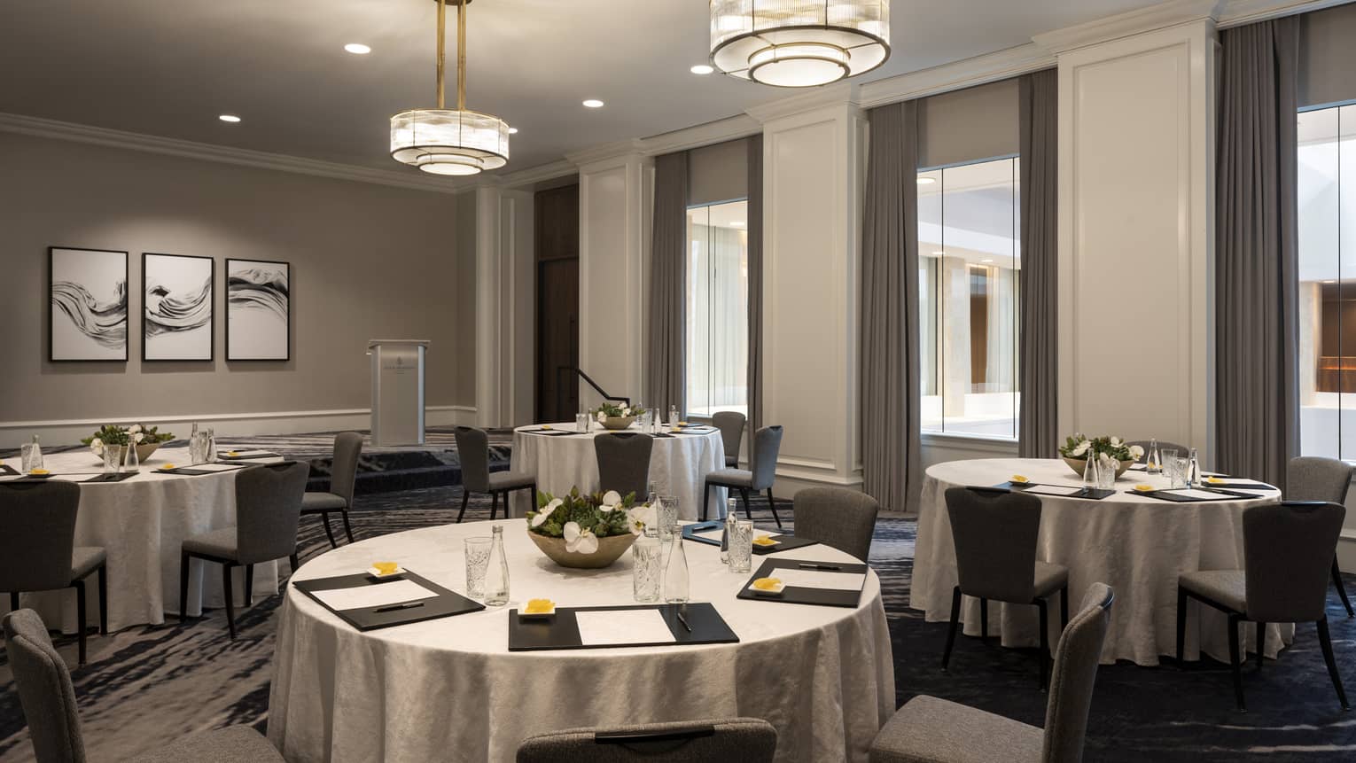 A room with large round tables covered in cloth. A menu is placed at each seat, while a flower arrangement is at the center of each table.