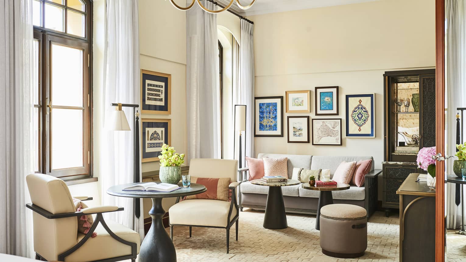 Hotel suite's living area, featuring a round table with two upholstered chairs, sofa, two coffee tables, colourful framed prints on the wall and two French windows