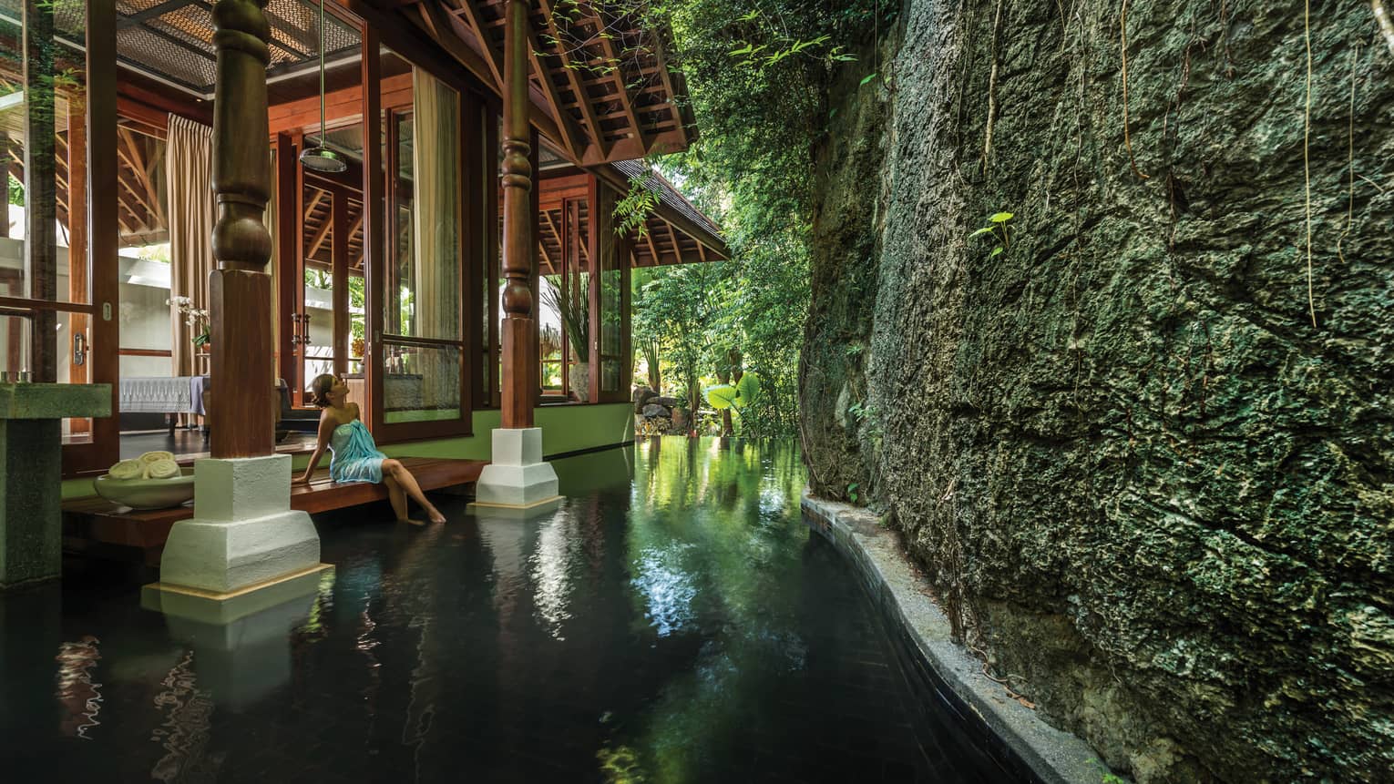 Woman in towel sits on spa steps with feet in pool below large rock wall