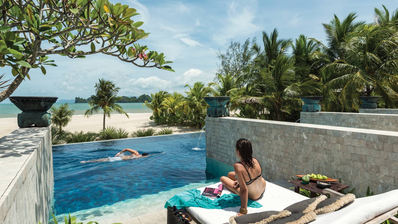 Back of woman in swimsuit on lounge chair, man swimming in pool below