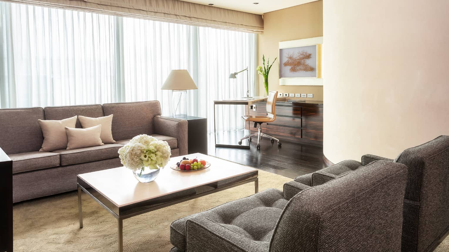 Living area with grey sofas, coffee table with flowers and fruit, work desk with a chair and floor-to-ceiling windows with curtains