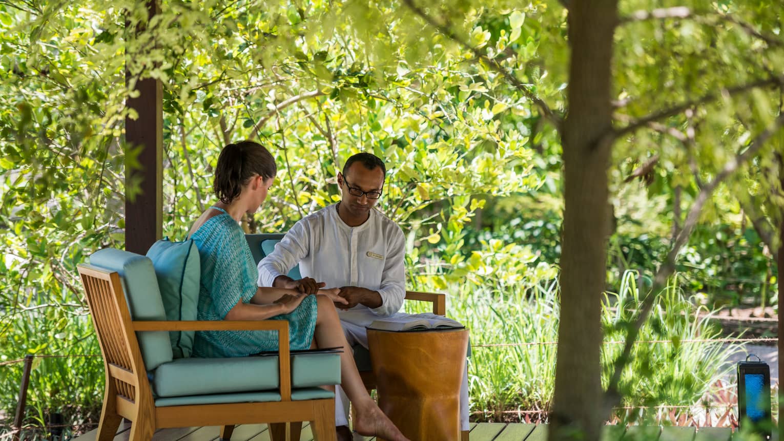 Spa staff places hand on woman's wrist by open book on plush blue chairs on patio pavilion