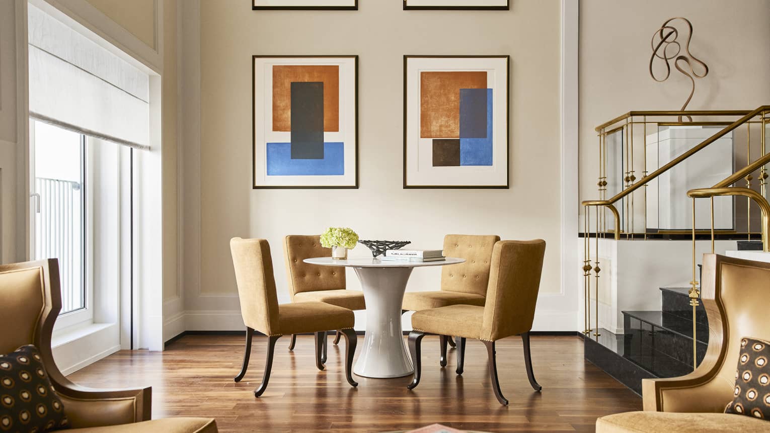Round dining table with four upholstered, tan chairs and four hanging paintings on wall