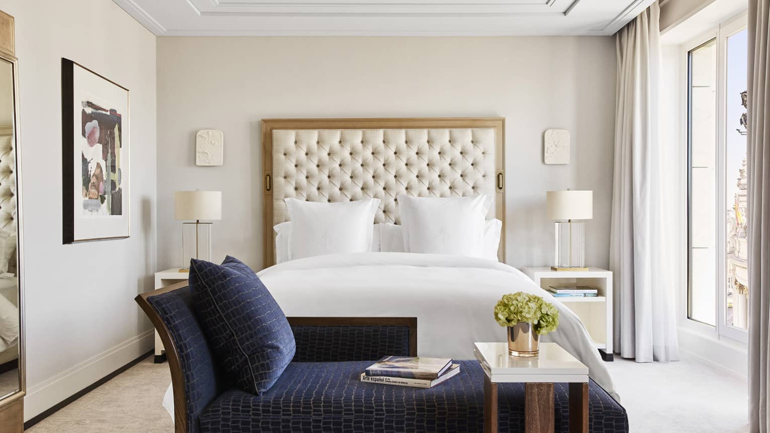 Bedroom with king bed, tufted headboard, dark blue chaise lounge at foot of bed, white ceiling molding