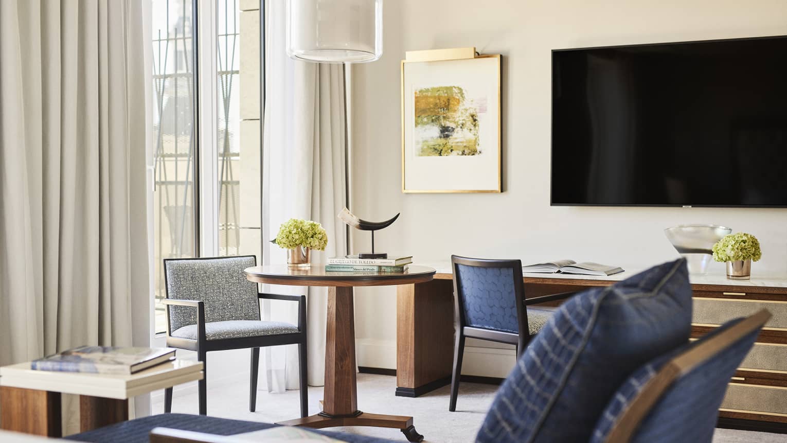 Luxury hotel room seating area with table with books, horn sculpture and flowers, chairs, blue chaise, TV, desk with decor, wall art and windows with curtains