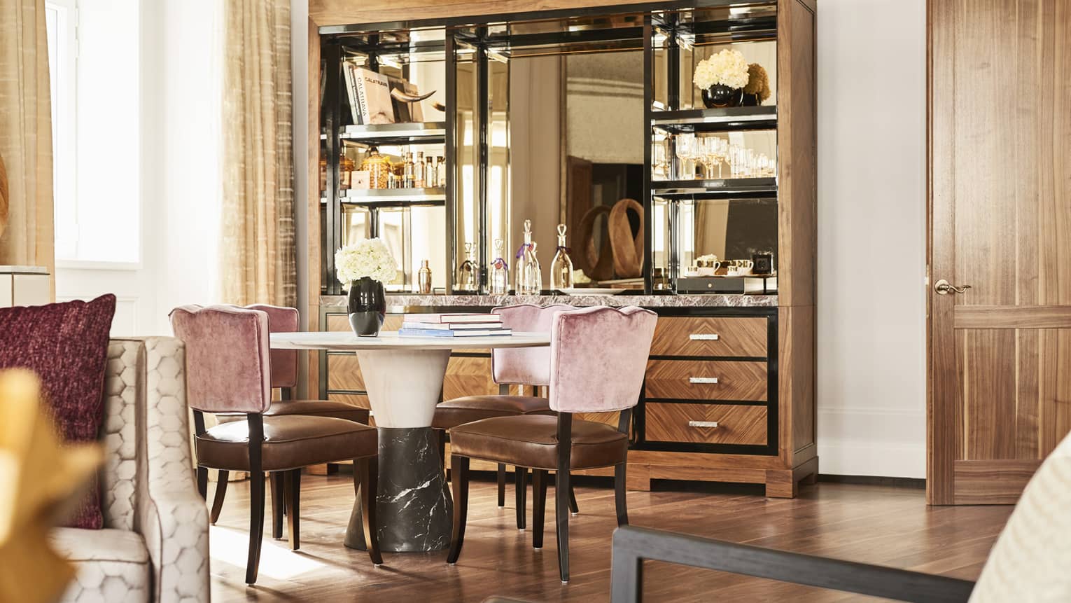 Luxury hotel suite dining area with round marble table, pink velvet and brown leather chairs, wood and mirrored cabinet displaying glassware and decor
