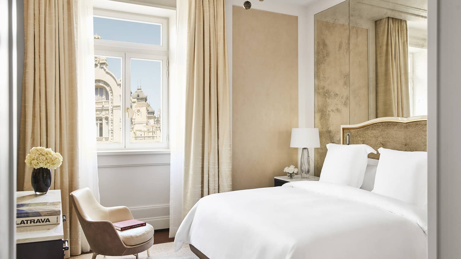 Bright luxury hotel bedroom with upholstered bed, mirrored wall panels, beige armchair, side table with flowers and window overlooking historic architecture
