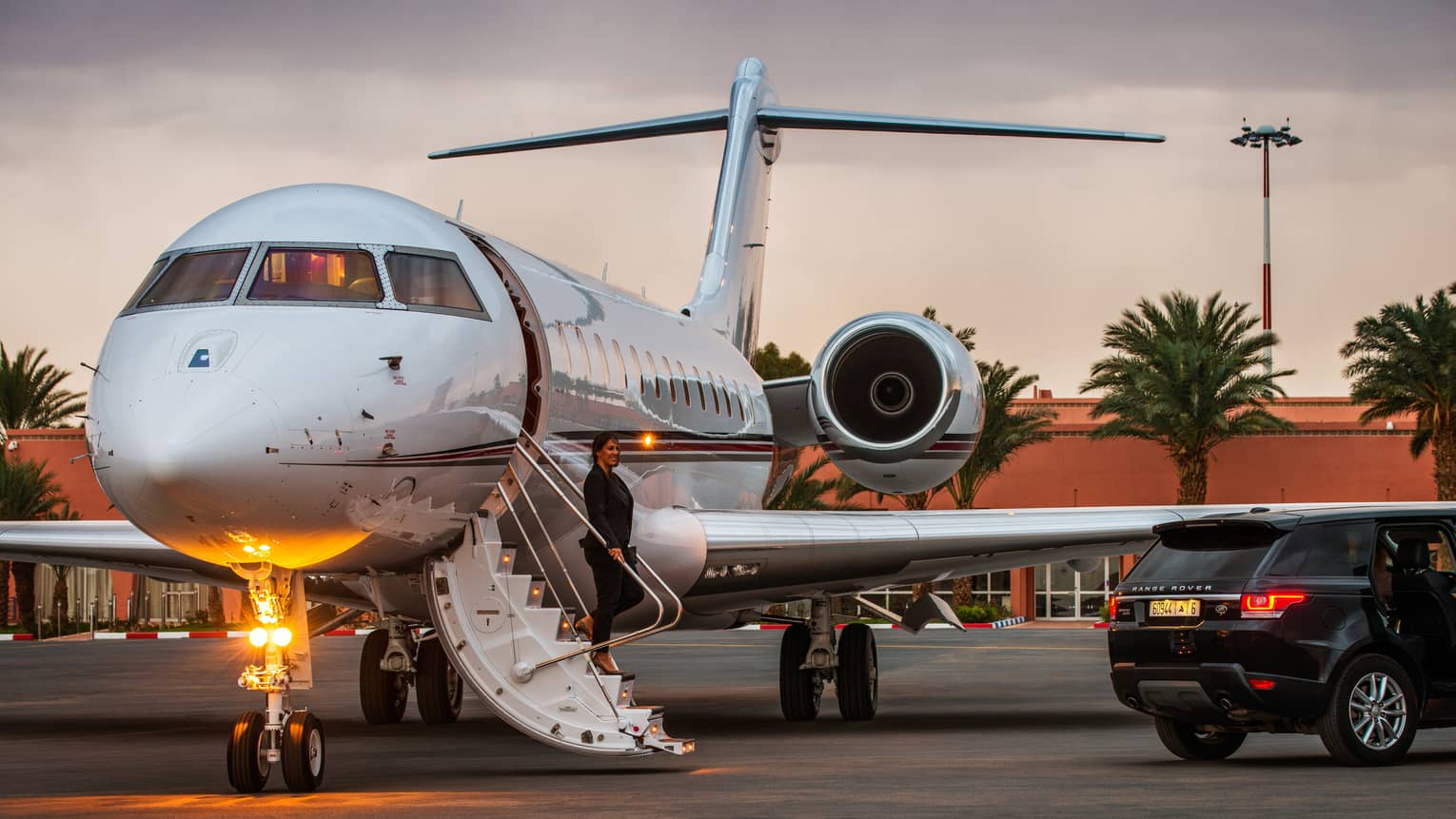 A woman stands on stairs of private plane, a black SUV is parked just outside