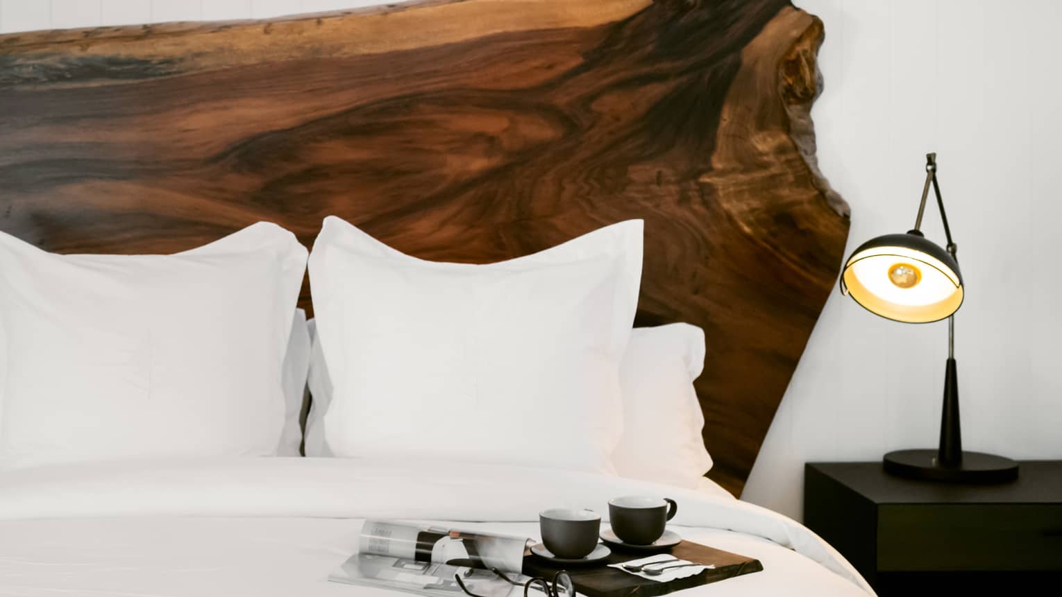 A hotel bed with a small black lamp next to it, a large natural wood headboard, and a tray of coffee resting on it.