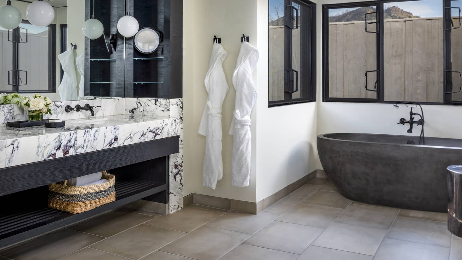 Luxury resort bathroom with a window-side dark freestanding bathtub, double marble vanity, hanging globe lights and two white bathrobes on the wall