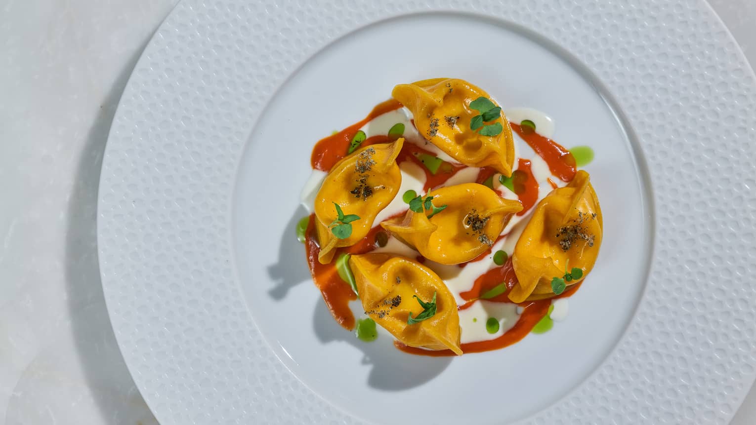 A white plate with a textured border holds five orange food items topped with green garnish and drizzled with a red sauce.