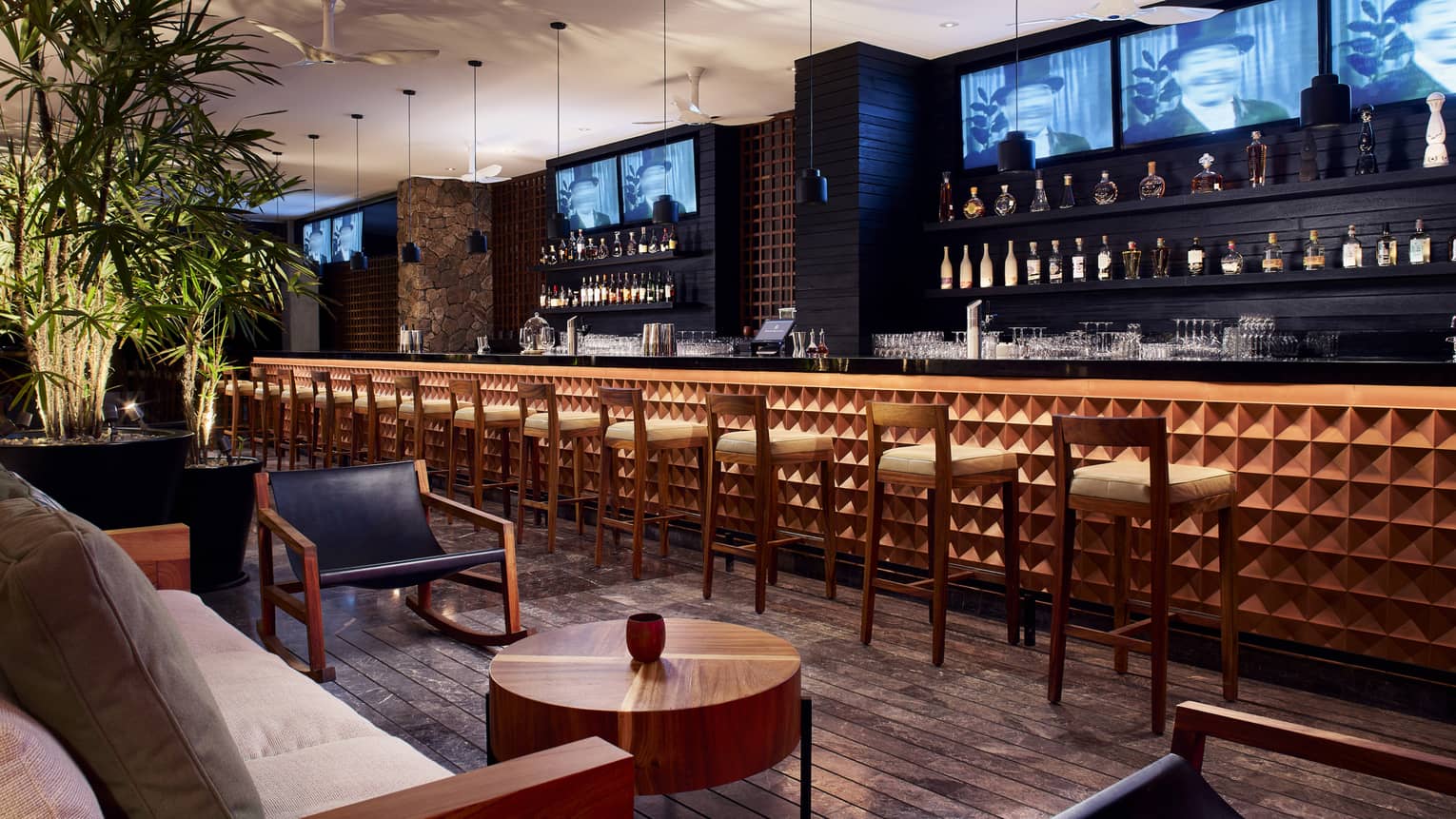 Chairs line a wooden bar with TV screens and shelves of bottles