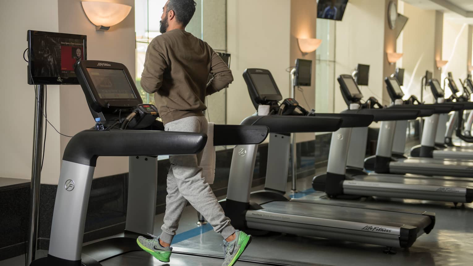 Back of man in sweatshirt and sweatpants as he runs on treadmill in gym next to row of cardio machines