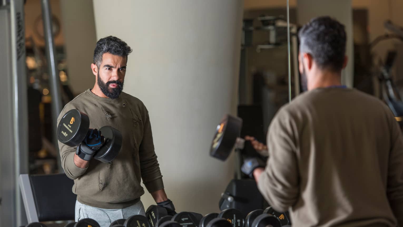 Man with beard wearing casual sweatshirt and sweatpants stands looks in gym mirror, lifts large hand weights
