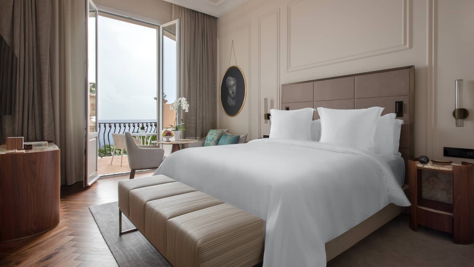 Hotel room with white king bed, tan leather bench, small balcony