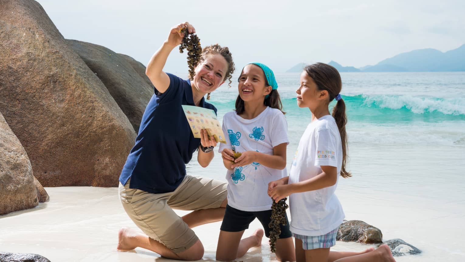 Woman kneels on white sand beach beside boulder, holds up seaweed to show two girls