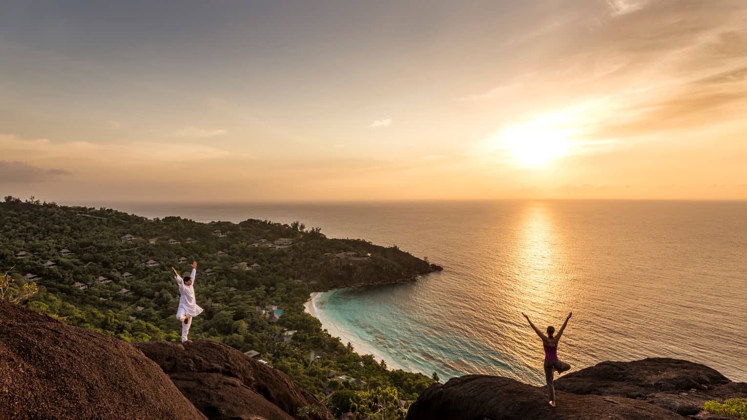 Woman and man stand apart on large boulders overlooking canopy of trees, ocean at sunrise 