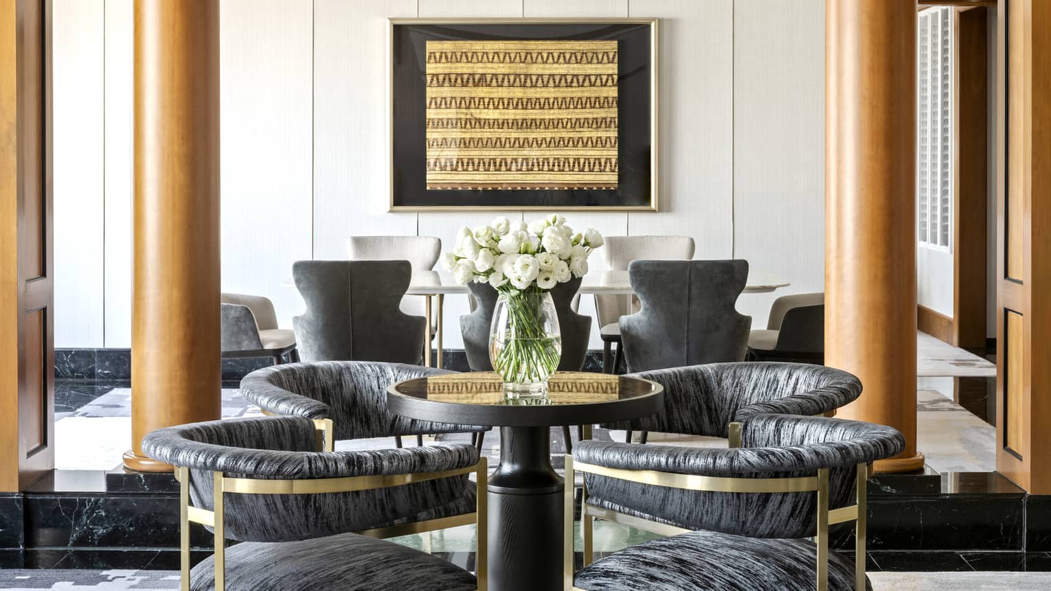 A stylish dining area featuring a small round table with modern cushioned chairs and a vase of white flowers. The space is accented by wooden columns and framed wall art.