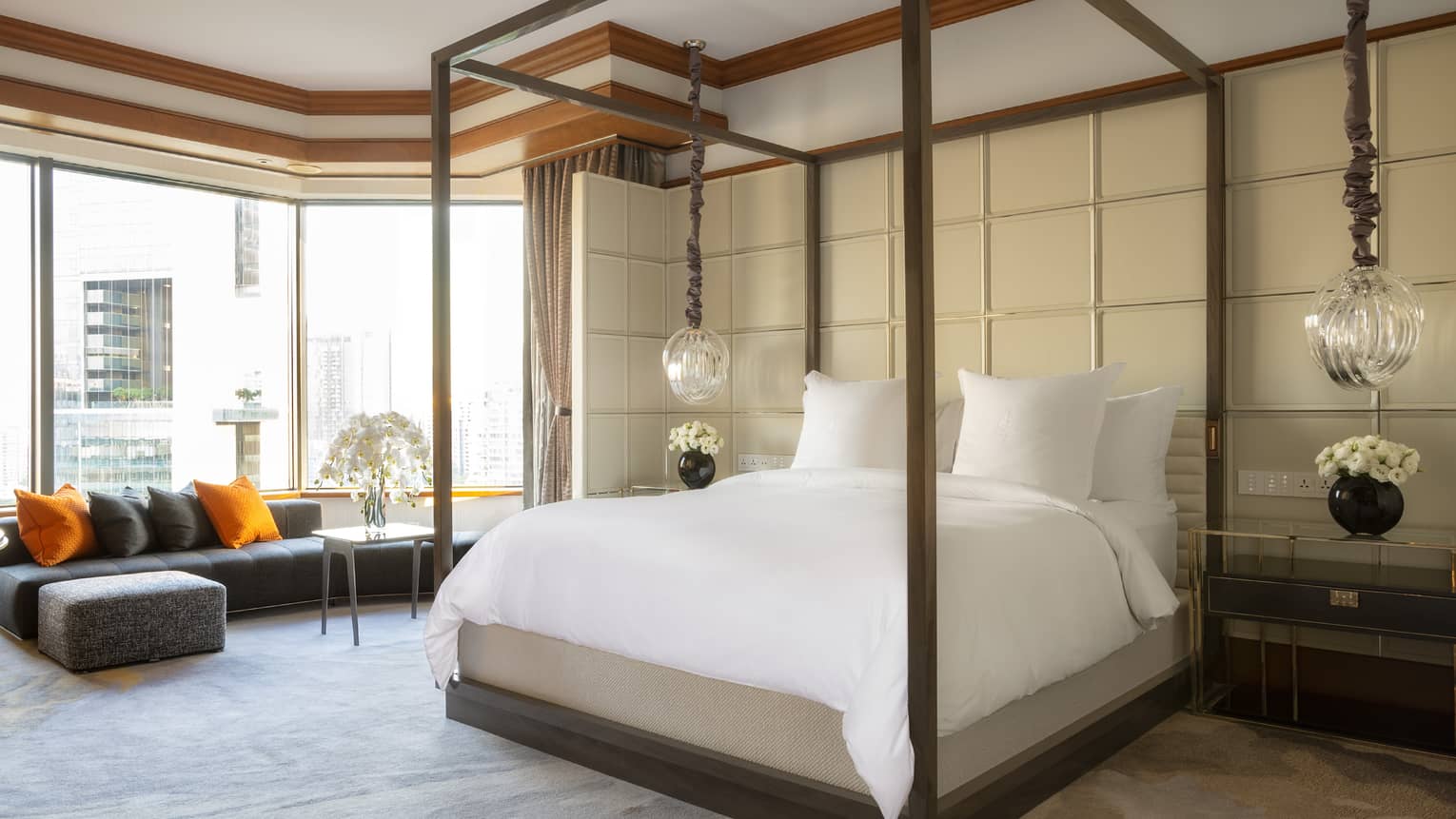A spacious bedroom with a four-poster bed, accented by hanging pendant lights and a cushioned seating area by large windows. The décor includes neutral tones with orange pillows.