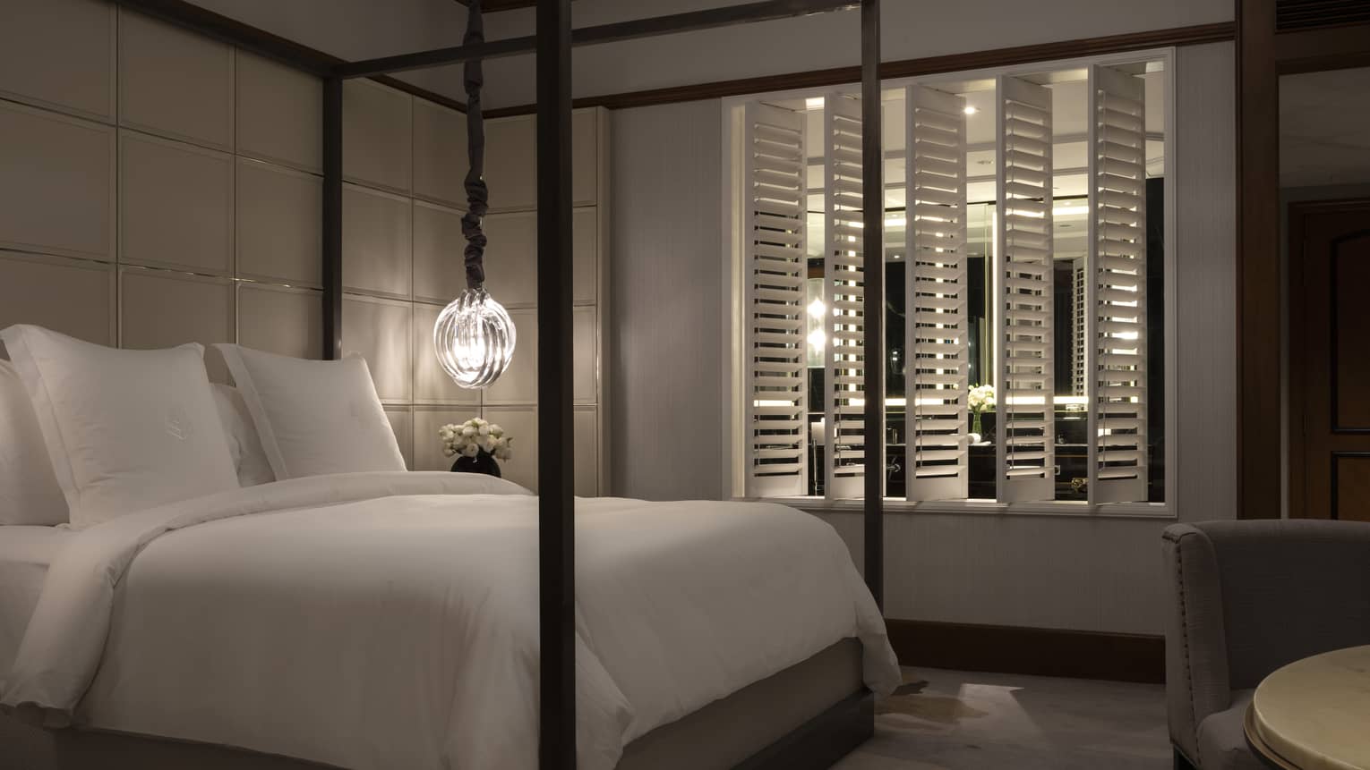 A dimly lit bedroom featuring a four-poster bed with white linens, hanging pendant light and shuttered window separating the bedroom from an adjacent room.