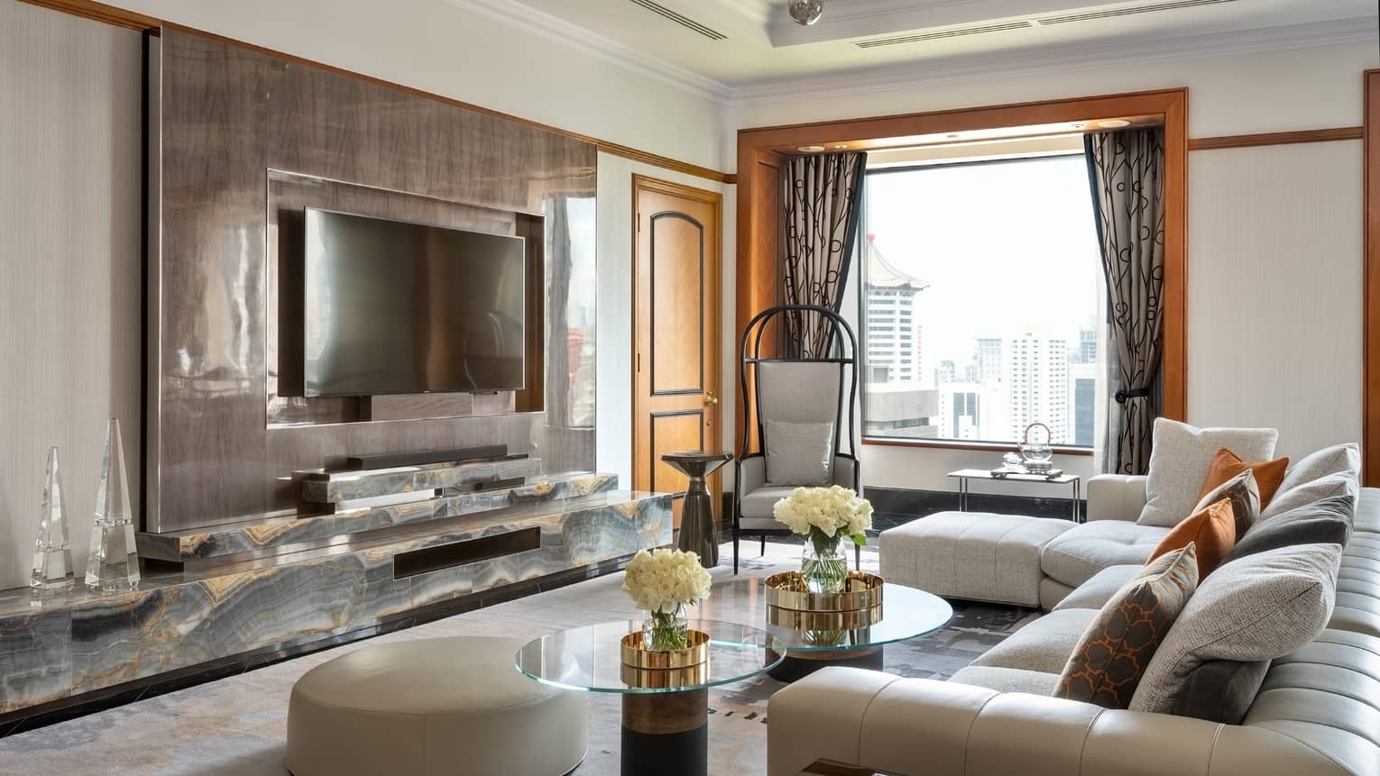 A modern living room with a large TV mounted on a polished wall, sectional sofa with orange and grey pillows, and glass-topped coffee tables. The room offers city views through large windows.