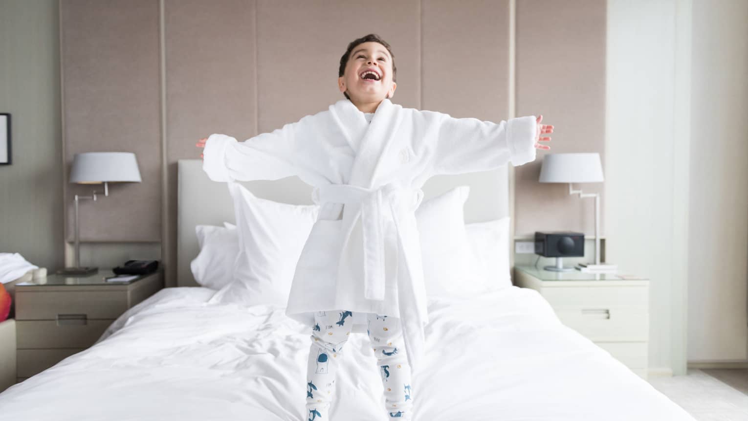 Smiling young boy wearing white bathrobe, pyjamas stands on hotel bed