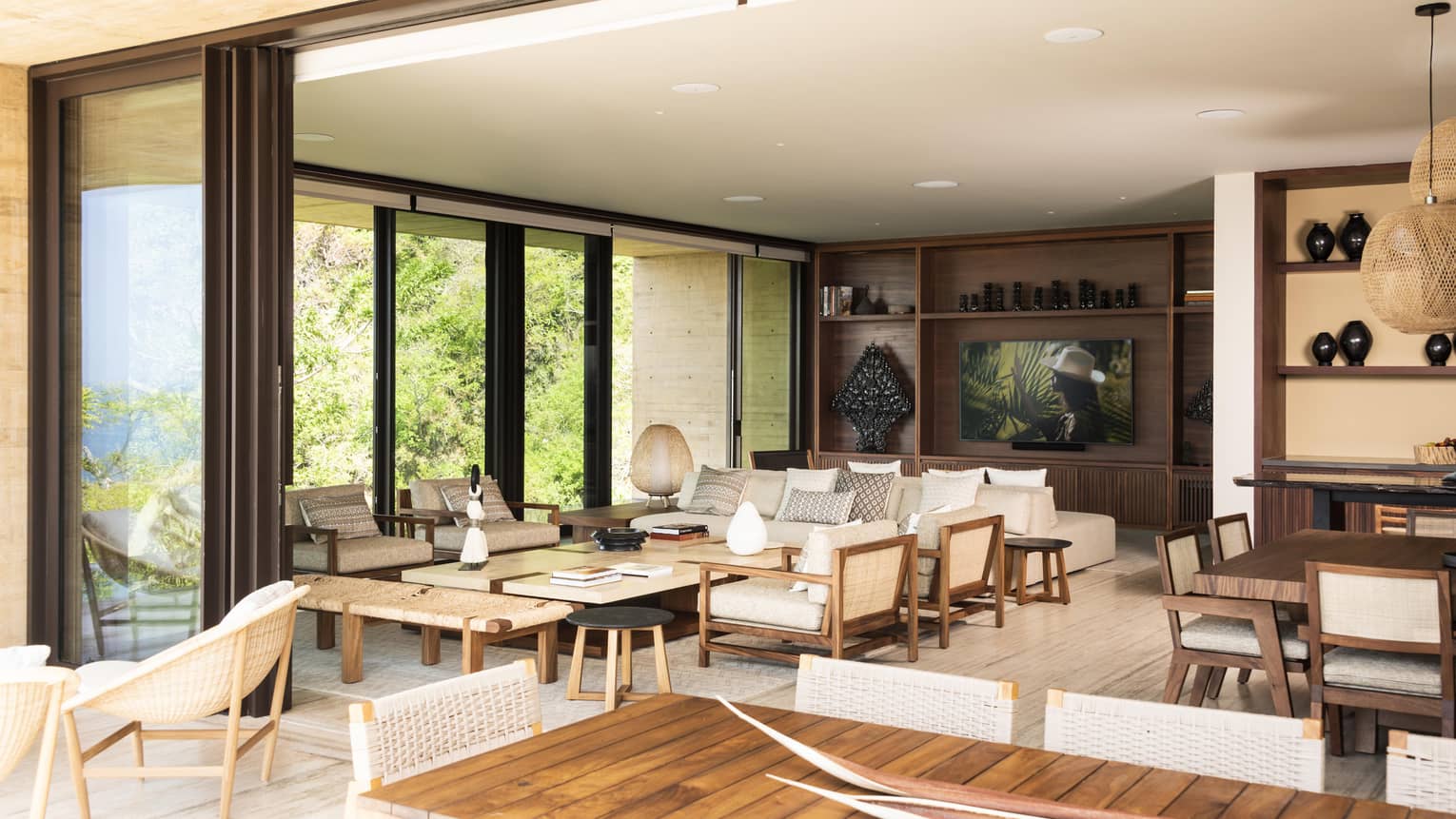 Spacious living area with floor-to-ceiling glass doors, wooden furniture and neutral decor. The room opens to an outdoor view with greenery, blending indoor and outdoor spaces.