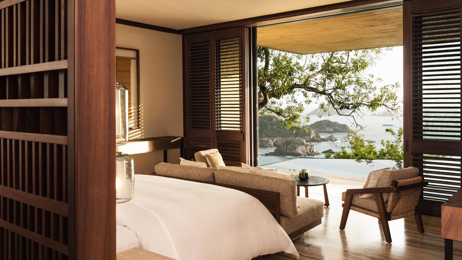 A serene hotel bedroom with wooden accents and a cozy seating area, opening to a terrace with breathtaking views of the ocean, rocky islands and lush greenery