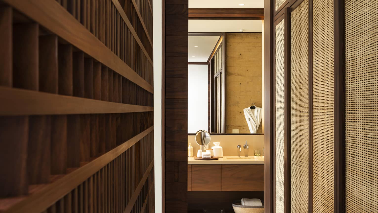 Hallway leading to a modern bathroom with wooden accents, featuring a double-sink vanity and a mirror. There are textured walls, and a bathrobe is hanging in the background.