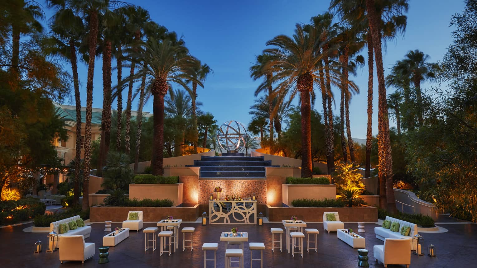 Palm trees over stone fountain, terrace seating area with lights at dusk